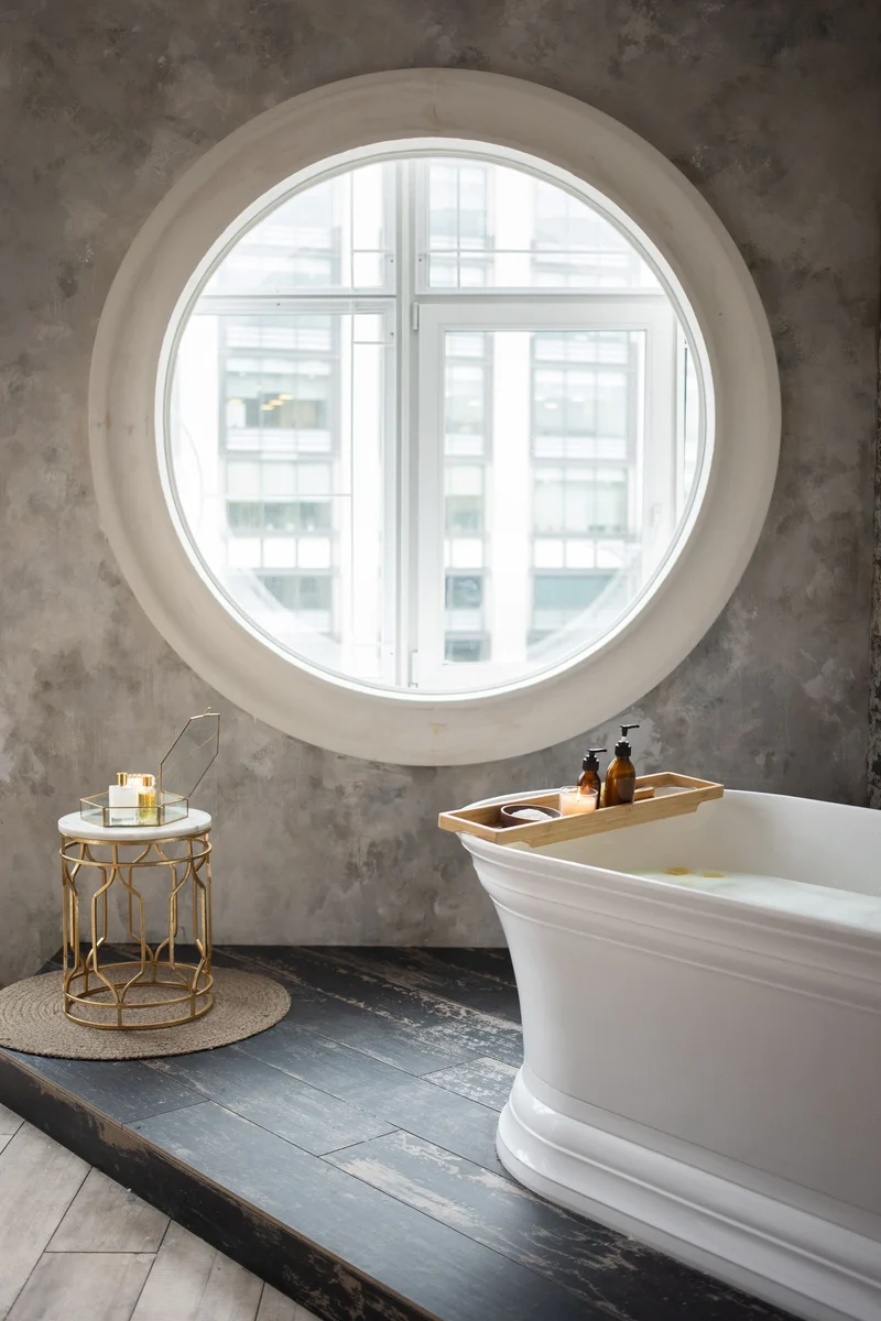 Salle de bains avec un mur gris à finition minérale, une fenêtre ronde et une baignoire blanche